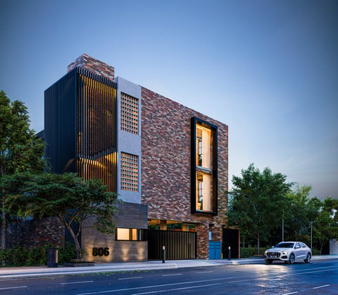 Modern multi-story brick building with vertical slat detailing, illuminated windows, and a white car parked outside, surrounded by trees against a clear sky