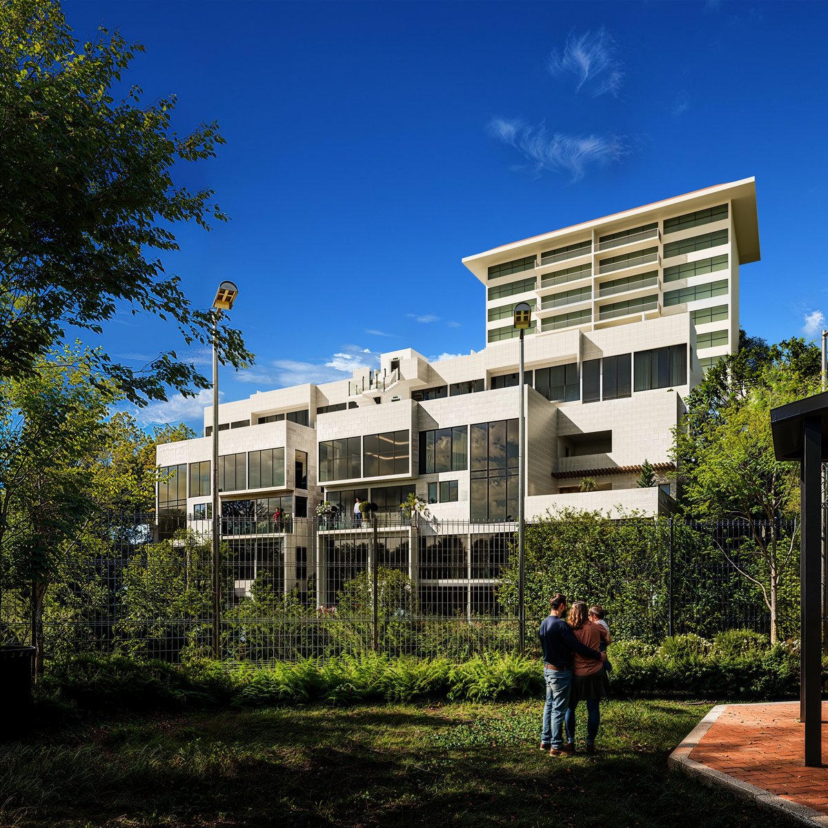 Modern white multi-story residential building with horizontal louvers, surrounded by green vegetation and blue sky, with two people standing in foreground
