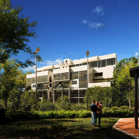 Modern white residential building with geometric design surrounded by green trees and landscaping, with two people viewing from terrace in foreground