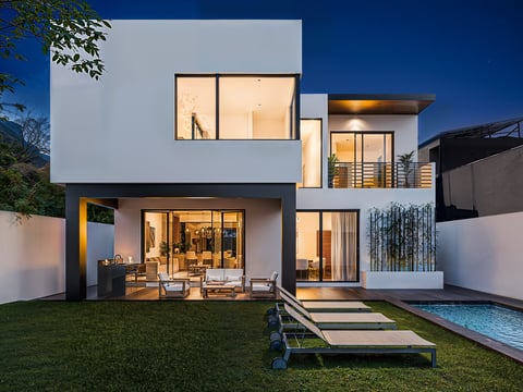 Modern white house with illuminated interior, manicured lawn, wooden lounge chairs, and pool at dusk