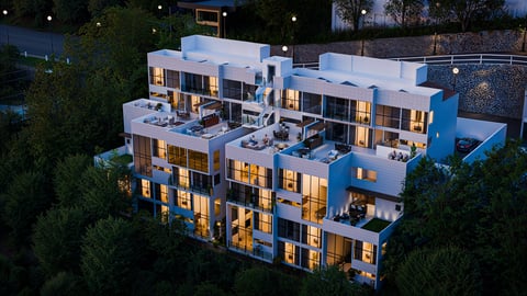 Modern apartment building at dusk with illuminated windows, white and blue exterior, surrounded by green landscaping